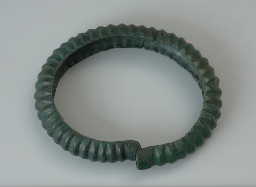 Bransoleta żebrowana - Ujęcie z góry. Bransoleta obręczowa wykonana z grubej brązowej blachy wygiętej w kształcie litery C. Owalna obręcz została ozdobiona głębokimi poprzecznymi żłobkami (żeberkami), natomiast jej końce, zachodzące na siebie, dekoruje motyw drobnej kratki. Powierzchnia zabytku pokryta jest zieloną patyną.