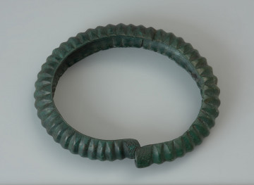 Bransoleta żebrowana - Ujęcie z góry. Bransoleta obręczowa wykonana z grubej brązowej blachy wygiętej w kształcie litery C. Owalna obręcz została ozdobiona głębokimi poprzecznymi żłobkami (żeberkami), natomiast jej końce, zachodzące na siebie, dekoruje motyw drobnej kratki. Powierzchnia zabytku pokryta jest zieloną patyną.