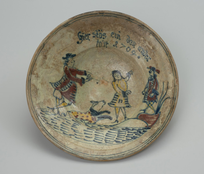 talerz - Ujęcie z przodu. Wykonany na kole garncarskim z gliny o jasnej barwie ( z mała ilością tlenków żelaza). Dno słabo wyodrębnione przechodzi w niewysoki korpus o szerokim wlewie z wyraźnym kołnierzem. Szkliwo w tonacji żółtoszarej tylko na licowej powierzchni naczynia. Temat ornamentu wskazuje z dużym prawdopodobieństwem na to, że talerz był wyrobem jednostkowym wykonanym na zamówienie. Malatura przedstawia scenę rodzajowa: kobieta w niekompletnym stroju stoi pomiędzy dwoma mężczyznami. Jedna z tych postaci (większa) wyciąga ręce ku kobiecie , obok niej pies. Ponad postaciami , na kołnierzu, umieszczono napis: 