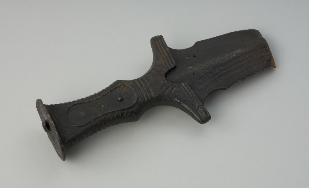 broń, miecz - Ujęcie w poziomie tyłu skosem w prawą stronę. Brązowy miecz z czworokatną, dekorowaną żłobieniami rękojeścią, głowicą w formie owalnej tarczki oraz jelcem ze skrzydełkami i półkolistym wycięciem.