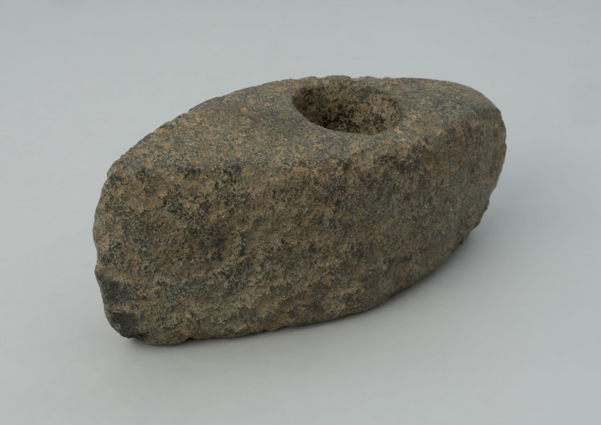 Topór kamienny - Ujęcie z przodu skosem w prawą stronę. Topór o wydłużonej części tylnej zakończonej wąskim obuchem, z wywierconych otworem przesuniętym ku tyłowi. Przekrój poprzeczny czworokątny wysoki. W w rzucie bocznym trapezowaty z lekko skośnym ostrzem i lekko łukowatym obuchem. Przy otworze, na stronie górnej, ślady wykucia płytkiej miseczki pod wiercenie. Powierzchnia zerodowana ze śladami szlifowania i puncowania. Ostrze wtórnie uszkodzone