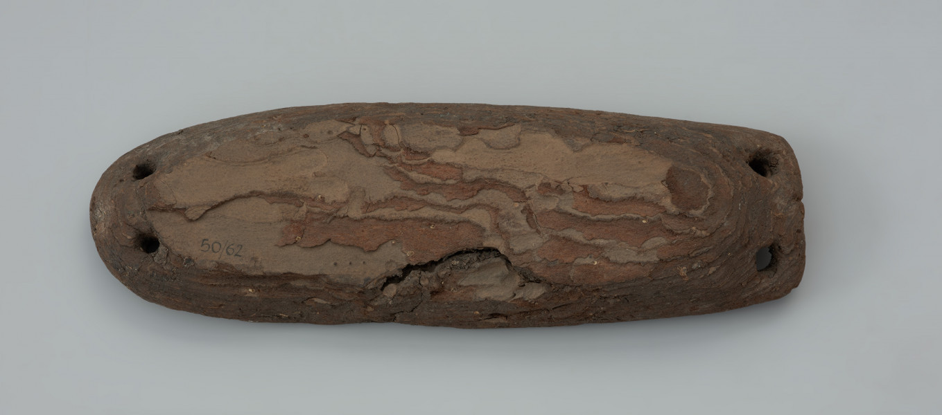 pływak - Ujęcie z góry przodu pływaka. Przedmiot wykonano z kawałka kory pochodzącej najprawdopodobniej z drzewa sosnowego. Zabytek ma wydłużony, owalny kształt o zaokrąglonym oraz ściętym końcu. Przedmiot uformowano za pomocą noża. Grzbiet oraz krawędzie zostały staranie ostrugane, natomiast dolna część pływaka jest naturalnie wklęsła odpowiadająca negatywowi pnia drzewa, z którego zdarto korę. Na obydwóch końcach znajdują się po dwa owalne otwory wywiercone czubkiem noża lub świdrem. Służyły one do mocowania lin połączonych bezpośrednio do sieci.