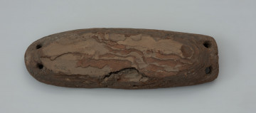 pływak - Ujęcie z góry przodu pływaka. Przedmiot wykonano z kawałka kory pochodzącej najprawdopodobniej z drzewa sosnowego. Zabytek ma wydłużony, owalny kształt o zaokrąglonym oraz ściętym końcu. Przedmiot uformowano za pomocą noża. Grzbiet oraz krawędzie zostały staranie ostrugane, natomiast dolna część pływaka jest naturalnie wklęsła odpowiadająca negatywowi pnia drzewa, z którego zdarto korę. Na obydwóch końcach znajdują się po dwa owalne otwory wywiercone czubkiem noża lub świdrem. Służyły one do mocowania lin połączonych bezpośrednio do sieci.