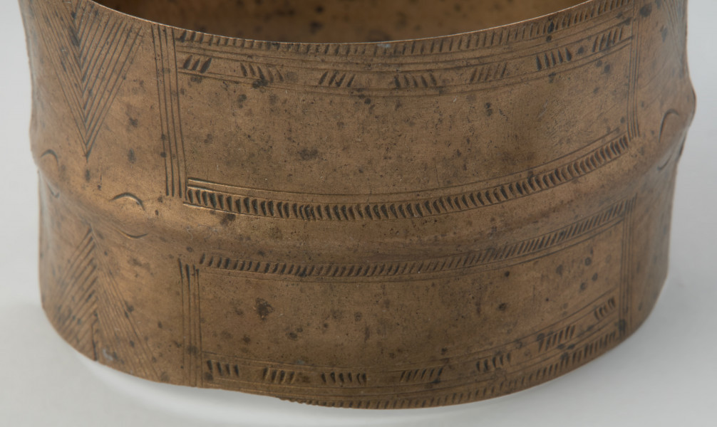 bransoleta taśmowata - Powiększenie detalu zdobień bransolety z przodu. Bransoleta otwarta wykonana z wąskiej, dość grubej taśmy brązowej o przekroju płasko-wypukłym. Końce obręczy są ścięte prosto, a jej powierzchnię pokrywa ornament ryty złożony z grup ukośnych i pionowych kresek, poprzedzielanych motywem jodełki.