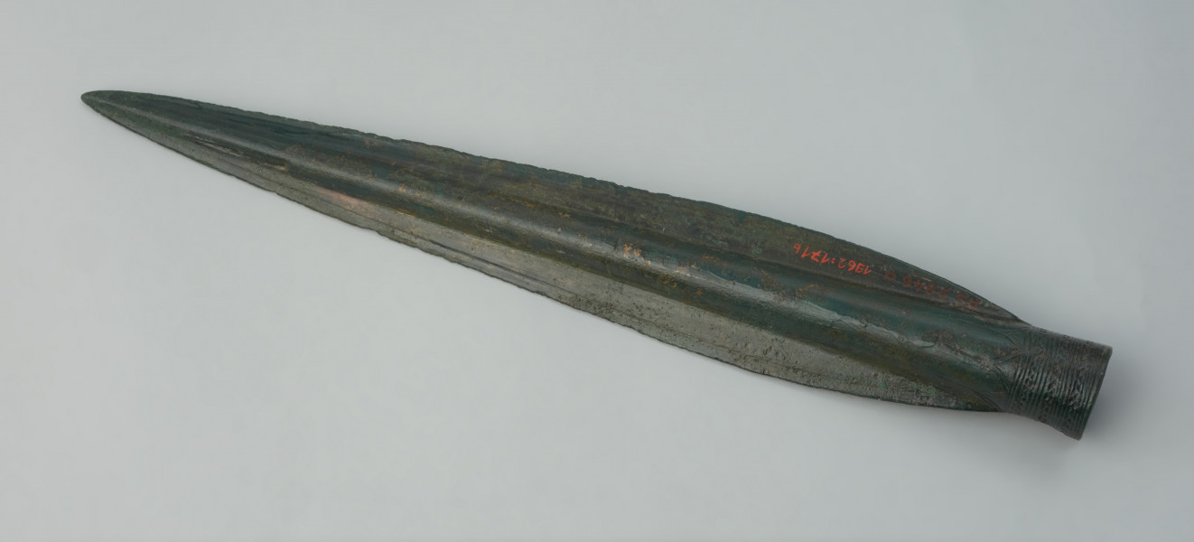 Grot oszczepu lub włóczni typu Ullerslev - Ujęcie w poziomie tyłu grotu. Długi, smukły grot o któtkiej tulejce, wzmocniony żeberkiem. Tulejka zdobiona jest przy wylocie trzema pasmami zygzakowymi, poprzedzielanymi pasmami dookolnych linii, całość wieńczą trójkąty. Zabytek pokrywa zielona patyna.