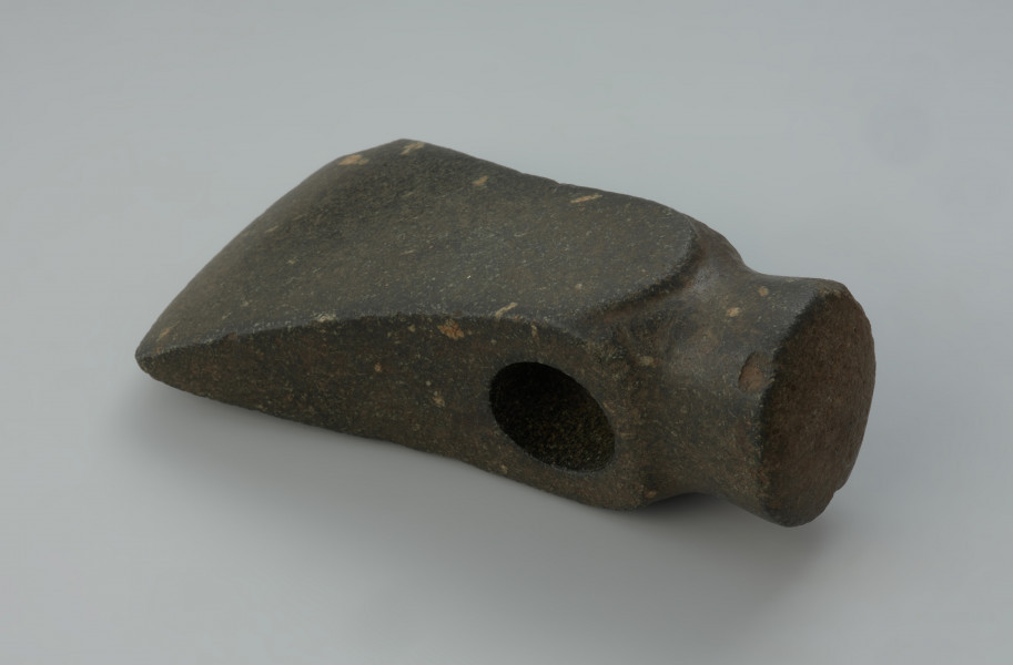 Miniaturowy kamienny toporek. - Ujęcie w poziomie boku toporka. Miniaturowy kamienny toporek z czopowatym obuchem oraz o korpusie lekko rozszerzającym się w kierunku ostrza.