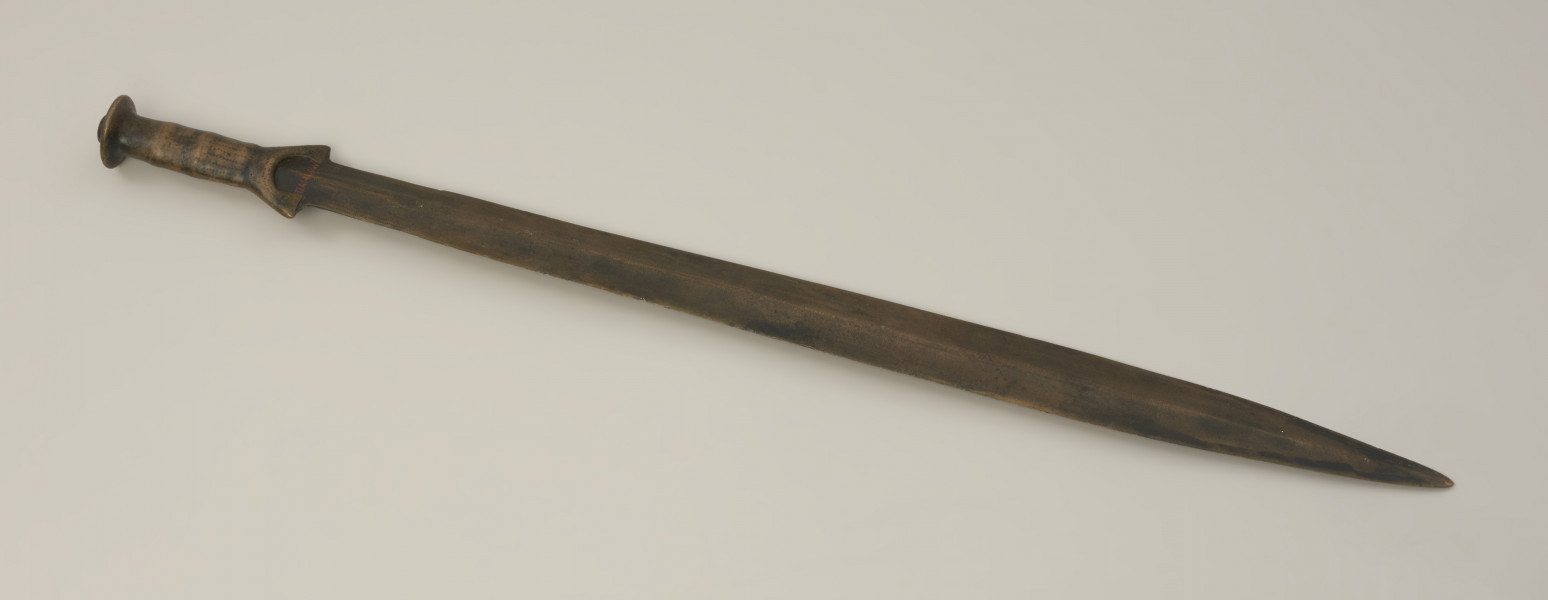 broń - Ujęcie z tyłu. Brązowy miecz z rękojeścią zakończoną tarczką oraz szeroką głownią. Całość jest bogato zdobiona ornamentem punktowym oraz nacięciami.
