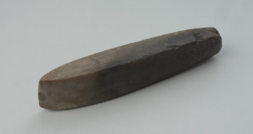 Dłuto krzemienne - Ujęcie z przodu skosem w lewą stronę. Dłuto krzemienne, czworościenne, trapezowate, o tępym obuchu. Powierzchnia szlifowana, przy ostrzu i w stronę obucha polerowana do połysku, a na obuchu negatywowa. Krzemień kredowy o barwie od jasno- do ciemnoszarej