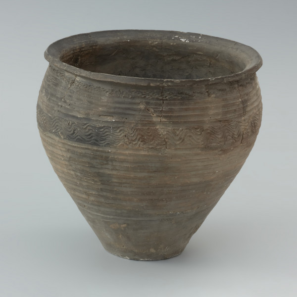 naczynie gliniane - Ujęcie z przodu. Garnek o baniastej formie z charakterystycznie wychyloną na zewnątrz krawędzią wylewu, obtoczony w całości na kole garncarskim. Brzusiec garnka znajduje się w 2/3 wysokości. Powierzchnia glinianego pojemnika przyozdobiona jest ornamentem wielowątkowym skośnych żłobków, dookolnych linii rytych, linii falistych oraz ponownych linii rytych. Fakturę naczynia tworzy dobrze wyprofilowana, gładka powierzchnia. Garnek wylepiono z glinianych wałeczków z wcześniej przygotowanej masy garncarskiej zawierającej domieszkę drobno i średnioziarnistego tłucznia - piasku i granitu.