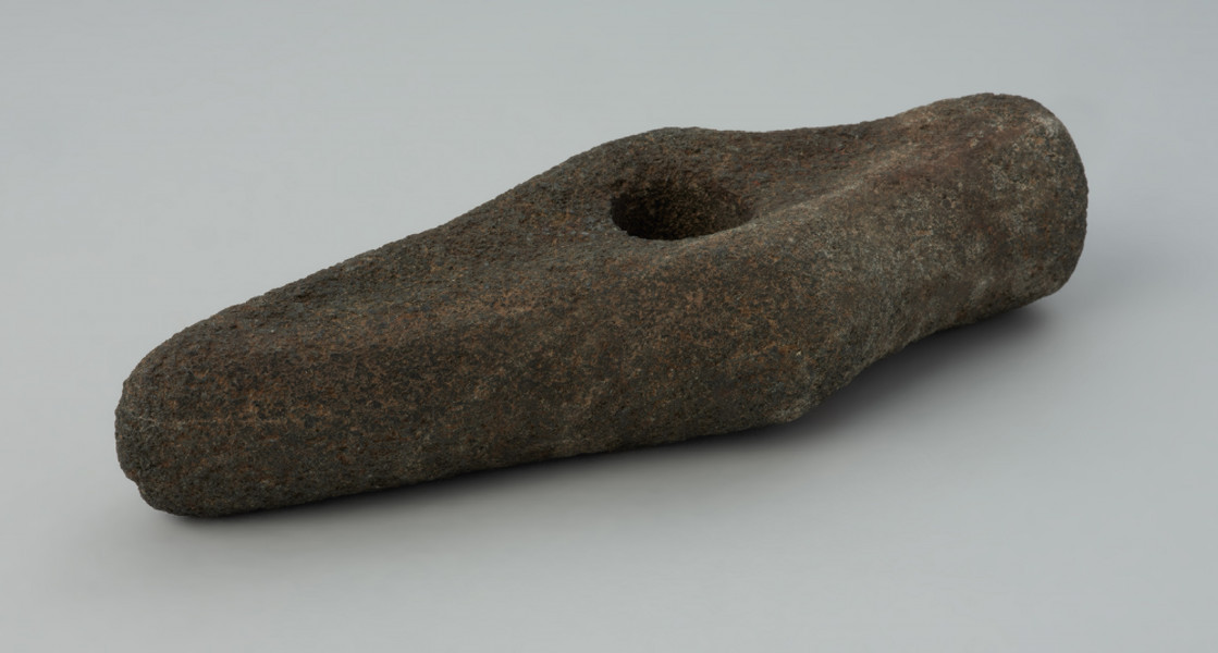 Topór kamienny - Ujęcie z przodu skosem w lewą stronę; Topór kamienny z wklęsłymi powierzchniami górną i dolną oraz lekko wypukłymi powierzchniami bocznymi. Ostrze proste. Obuch tępym o zarysie kolistym. Korpus prosty w rzucie bocznym, w rzucie z góry z poszerzeniem przy otworze. Otwór kolisty, cylindryczny. Powierzchnia zerodowana, pierwotnie gładzona.