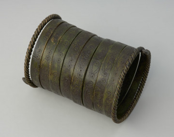 bransoleta spiralna typu kujawskiego - Ujęcie boku skosem w lewo. Jedenastozwojowa bransoleta spiralna wykonana z wąskiej, brązowej taśmy. Zwoje ornamentowane są tzw. motywem oczkowym oraz punktowanym zygzakiem. Końce bransolety ozdobiono tordowaniem.