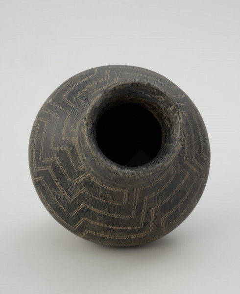 naczynie gliniane - Ujęcie z góry. Gliniane naczynie wąskootworowe o flaszowatej formie z wysoką cylindryczną szyjką. Powierzchnia jest wyświecana, barwy ciemnobrunatnej. Brzusiec jest dwustożkowaty, zdobiony techniką kółka zębatego motywem meandra schodkowego, a w dolnej partii pionowymi liniami. Szyjkę obiegają u podstawy dwie listy plastyczne.