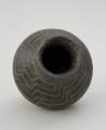 naczynie gliniane - Ujęcie z góry. Gliniane naczynie wąskootworowe o flaszowatej formie z wysoką cylindryczną szyjką. Powierzchnia jest wyświecana, barwy ciemnobrunatnej. Brzusiec jest dwustożkowaty, zdobiony techniką kółka zębatego motywem meandra schodkowego, a w dolnej partii pionowymi liniami. Szyjkę obiegają u podstawy dwie listy plastyczne.