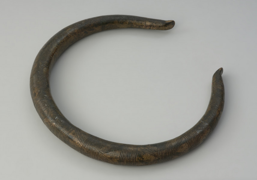 Brązowy naszyjnik - Ujęcie z góry skosem w lewo naszyjnika. Brązowy naszyjnik wykonany z grubego okrągłego pręta. Jego zakończenia są odgięte na zewnątrz. Zdobiony jest poprzecznymi kreskami, trójkątami oraz łukami.