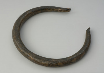 Brązowy naszyjnik - Ujęcie z góry skosem w lewo naszyjnika. Brązowy naszyjnik wykonany z grubego okrągłego pręta. Jego zakończenia są odgięte na zewnątrz. Zdobiony jest poprzecznymi kreskami, trójkątami oraz łukami.