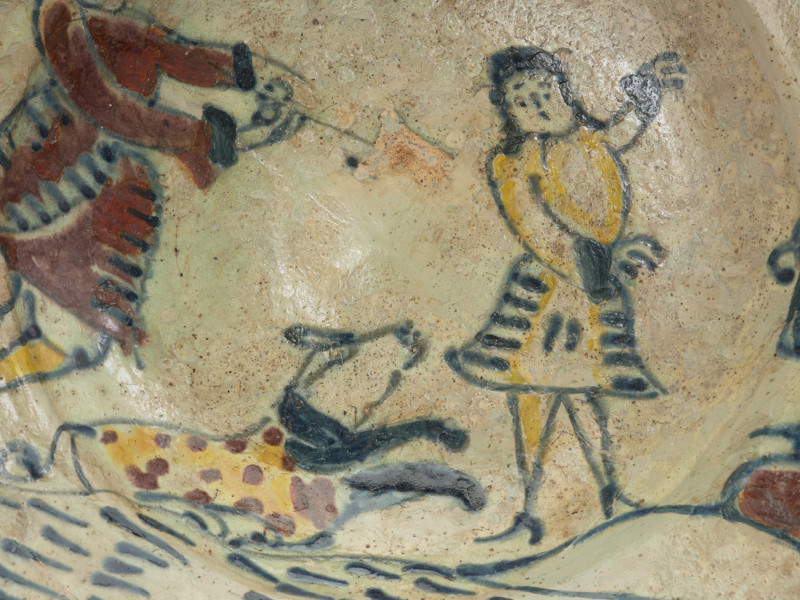 talerz - Ujęcie drugiego fragmentu. Wykonany na kole garncarskim z gliny o jasnej barwie ( z mała ilością tlenków żelaza). Dno słabo wyodrębnione przechodzi w niewysoki korpus o szerokim wlewie z wyraźnym kołnierzem. Szkliwo w tonacji żółtoszarej tylko na licowej powierzchni naczynia. Temat ornamentu wskazuje z dużym prawdopodobieństwem na to, że talerz był wyrobem jednostkowym wykonanym na zamówienie. Malatura przedstawia scenę rodzajowa: kobieta w niekompletnym stroju stoi pomiędzy dwoma mężczyznami. Jedna z tych postaci (większa) wyciąga ręce ku kobiecie , obok niej pies. Ponad postaciami , na kołnierzu, umieszczono napis: 