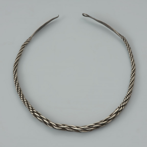 naszyjnik srebrny - Ujęcie przodu naszyjnika z góry. Srebrny naszyjnik spleciony z pięciu par drutów owiniętych wokół siebie, z końcami rozklepanymi w owalne, niezdobione płytki. Jedna z nich zakończona została haczykiem, końcówka drugiej jest ułamana. W części środkowej druty są skręcone luźniej, co powoduje, ze obręcz jest w tym miejscu grubsza, natomiast po obu stronach znajdują się fragmenty, gdzie splot jest nierówny.