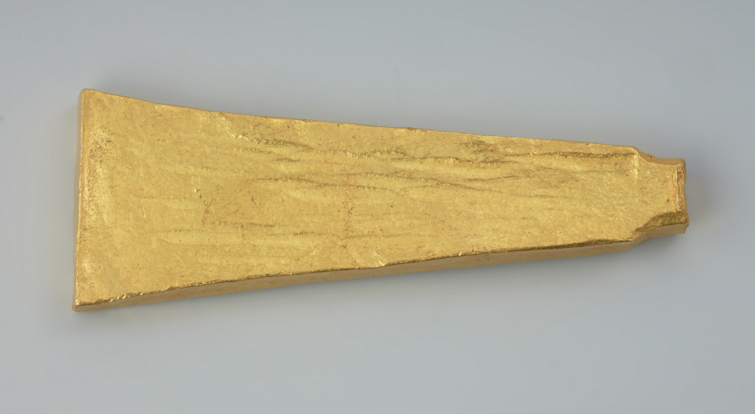 Sztabkowata płytka ze złota - Ujęcie z tyłu. Sztabkowata płytka ze złota w formie symetrycznego klina przechodzącego u szczytu w imitację owalnej w przekroju nasady lub ucha. Płytka jest zdobiona tylko na stronie wierzchniej.