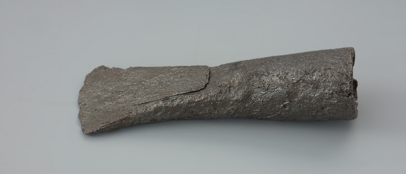 ciosła - Ujęcie z góry tyłu ciosła. Ciosła z okrągłą tulejkę do osadzania na trzonku. Tulejka jest lekko przewężona w części środkowej. Ostrze ciosły jest łukowate, stosunkowo krótkie, symetryczne. Po obydwóch stronach ostrza widoczne są ślady skuwania co najmniej dwóch odrębnych części materiału. Powierzchnia zabytku jest mocno skorodowana z licznymi ubytkami, zwłaszcza przy tulejce.