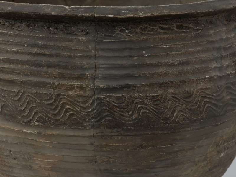 naczynie gliniane - Ujęcie górnego fragmentu lewej strony ścianki naczynia. Garnek o baniastej formie z charakterystycznie wychyloną na zewnątrz krawędzią wylewu, obtoczony w całości na kole garncarskim. Brzusiec garnka znajduje się w 2/3 wysokości. Powierzchnia glinianego pojemnika przyozdobiona jest ornamentem wielowątkowym skośnych żłobków, dookolnych linii rytych, linii falistych oraz ponownych linii rytych. Fakturę naczynia tworzy dobrze wyprofilowana, gładka powierzchnia. Garnek wylepiono z glinianych wałeczków z wcześniej przygotowanej masy garncarskiej zawierającej domieszkę drobno i średnioziarnistego tłucznia - piasku i granitu.