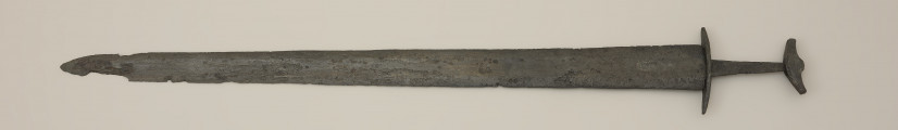 miecz dwusieczny - Ujęcie z przodu. Całkowicie zachowany, z niewielkimi ubytkami korozyjnymi w okolicach sztycha. Jego głownia jest najszersza przy jelcu i równomiernie zwężą się ku ostremu sztychowi. Jest zaopatrzona w szerokie zbrocze. Głownię wykonano w technice prętowej, poprzez zgrzanie stalowych nakładek ostrzowych z jednolitym rdzeniem. Trzpień mocno zwęża się ku głowicy. Jelec, soczewkowaty w rzucie z góry, jest długi i płaski, a jego końce są zakrzywione w kierunku głowni. Jednolita głowica ma kształt siodełkowaty. Jej dolna część wygięta jest ku górze. Na jej powierzchni zaznaczono symboliczny podział na podstawę i nakładkę.
