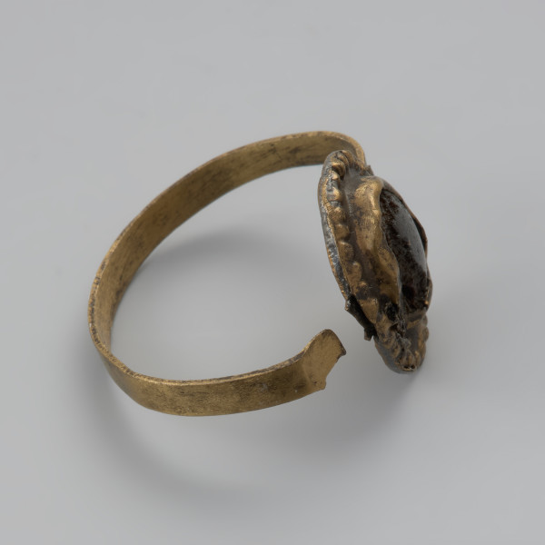 pierścionek z oczkiem - Ujęcie z góry lewej strony. Trzyczęściowy brązowy pierścionek z oczkiem, składający się z metalowej taśmy stanowiącej otok, metalowej tarczki pod oczko złożonej z owalnej płaskiej blaszki spodniej oraz dodatkowej taśmy tworzącej eliptyczne gniazdko. Jest ona zdobiona dookoła odciskami. Trzecim elementem, który uzupełnia oczko jest grafitowe szkiełko zachowane jedynie w części, przyklejone do spodniej tarczki i przytrzymywane przez krawędzie otaczającej je taśmy. Między blaszkami oczka pozostawione są dwie 