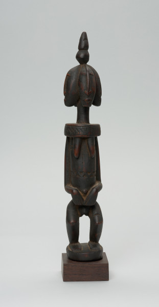 rzeźba - Ujęcie z przodu. Drewniana, rzeźbiona postać z wyraźnie zaznaczonymi piersiami, a także z czterema głowami. Każda głowa zwrócona jest w inna stronę. Na czubku głowy stożkowata bryła. Twarze zaznaczone bardzo symbolicznie, nos w kształcie strzałki. Ramiona od szyi oddzielone prostą linią. Ręce przylegające do tułowia, dłonie złożone na łonie/podbrzuszu. Nogi krótkie, podkurczone w kolanach. Na brzuchu, na wysokości pępka dekoracyjne trójkątne nacięcia.