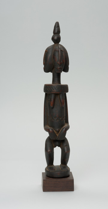 rzeźba - Ujęcie z przodu. Drewniana, rzeźbiona postać z wyraźnie zaznaczonymi piersiami, a także z czterema głowami. Każda głowa zwrócona jest w inna stronę. Na czubku głowy stożkowata bryła. Twarze zaznaczone bardzo symbolicznie, nos w kształcie strzałki. Ramiona od szyi oddzielone prostą linią. Ręce przylegające do tułowia, dłonie złożone na łonie/podbrzuszu. Nogi krótkie, podkurczone w kolanach. Na brzuchu, na wysokości pępka dekoracyjne trójkątne nacięcia.