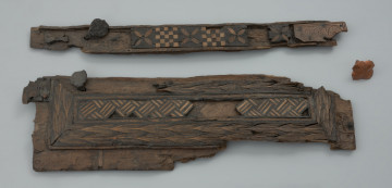 szkatułka intarsjowana - Ujęcie z góry. Wieko szkatułki z drewna bukowego, intarsjowana drewnem dębowym i cisowym. Ornamentykę szkatułki tworzą trzy odmienne pasma intarsji. Część centralną, prostokątną, wydzieloną obramowaniem zajmuje motyw ukośnie ułożonych kwadratowych płycin uzupełnianych trójkątami. Każdą płycinę wypełniają wąskie listewki, naprzemiennie ciemnej dębiny i jasnego cisu. Całość sprawia wrażenie grubej plecionki. Bezpośrednio do obramowania pasma centralnego przylega dekoracja w postaci dwóch rzędów kłosów z jasnego cisu. Przybrzeżną część ścianki otacza kompozycja złożona z naprzemiennie umieszczanych motywów: szachownic o 25 polach i czterolistnych rozet z jasnego drewna, precyzyjnie wkomponowanych w soczewkowate wycięcia w płycinach z ciemniejszej dębiny.