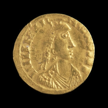 MNS/A/22203/1 złota moneta - Ujęcie awersu. Złoty solid Walentyniana III. Powierzchnie są starte. Moneta jest zapewne ostrogockim naśladownictwem wybitym w Rawennie. Na awersie widnieje popiersie cesarza w diademie i udrapowanej szacie zwrócone twarzą w prawa stronę. Wokół nieczytelny napis. Na rewersie postać cesarza stojąca przodem z krzyżem w prawej ręce i wyobrażeniem Victorii w lewej. Prawa stopa spoczywa na postaci węża z ludzką głową. Po obu stronach postaci cesarza znajdują się starte litery R i V, a pod stopami napis COMOB. W otoku monety figuruje napis VICTORIA AUGGG.