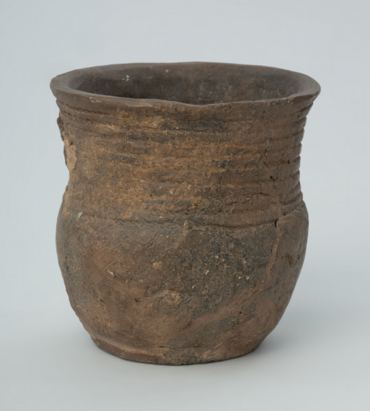 Naczynie gliniane - Ujęcie z przodu; Puchar o kształcie esowatym, być może pierwotnie zaopatrzony w ucho, zdobiony na szyjce dookolnym pasmem złożonym z 10 rzędów ornamentu odcisków sznura. Naczynie zrekonstruowane na podstawie drobnych fragmentów wylewu, szyjki, brzuśca i dna, uzupełnione barwionym gipsem. Powierzchnia szarobrunatna, z powierzchnią niedokładnie wyrównaną. W masie, na powierzchni widoczna domieszka mineralna.