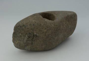 topór, broń obuchowa - Ujęcie tyłu; Topór pięcioboczny o wydłużonej części obuchowej z wąskim obuchem. Otwór wiercony dwustronnie, umieszczonym około połowy długości. Przekrój poprzeczny w formie wysokiego czworokąta. W rzucie bocznym trapezowaty, z wypukłą krawędzią górną. Powierzchnia szlifowana, strona górna zeszlifowana mocniej ma wyraźnie zaznaczone krawędzie. Na jednym z boków w części obuchowej widoczne ślady puncowania (obróbki przed szlifowaniem) i rozległego, zbyt głębokiego odbicia. Ostrze ma przebieg falisty z widocznymi obustronnymi, drobnymi odbiciami sugerującymi zużycie i prawdopodobnie przygotowanie do ostrzenia. Na powierzchni bocznej widoczne ślady wtórnych, znacznie świeższych uszkodzeń.