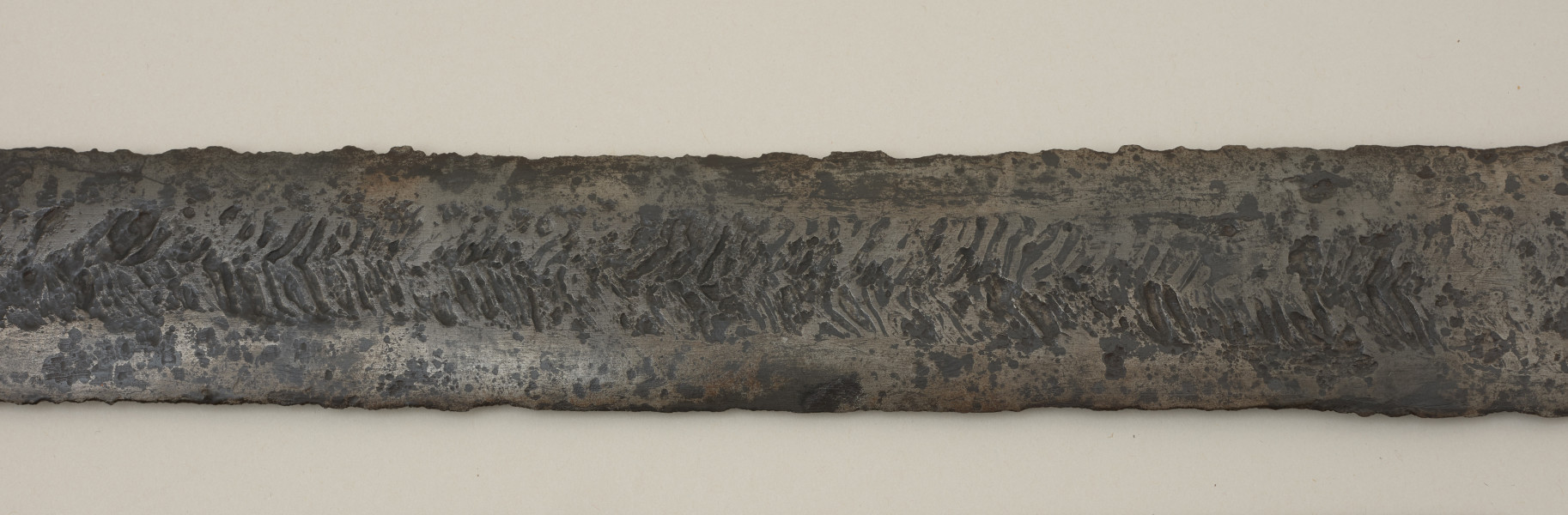 broń; miecz dwusieczny - Ujęcie głowni miecza. Całkowicie zachowany. Dwusieczna głownia jest szeroka i masywna, o mniej więcej jednakowej szerokości na całej długości, przy zastawie ma przekrój sześcioboczny. Sztych jest zaokrąglony. Na obu płazach głowni widoczny wzór w postaci „jodełki” świadczący o wieloelementowej jej konstrukcji. Rdzeń został wykonany z dwóch skręconych prętów, nisko- i wysokowęglowego, tworzących jądro, z dodanymi po obu stronach nakładkami ostrzy. Trzpień miecza jest wąski i lekko zwęża się ku głowicy. Jelec jest krótki i wysoki, o kształcie widzianym od góry zbliżonym do prostokąta o lekko wypukłych krótszych bokach. Głowica jest jednolita, zbliżona do trójkąta. Jelec i głowica są lite, wykute każde z pojedynczego kawałka metalu.