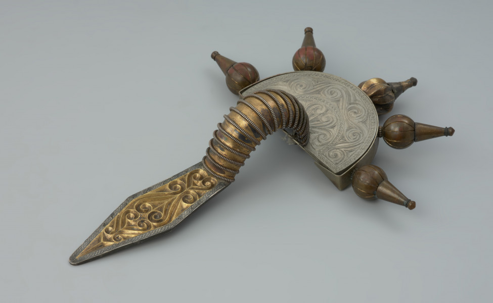 ozdoba; Srebrna, pozłacana fibula - Ujęcie z tyłu. Srebrna, pozłacana fibula z rombowato ukształtowaną długą nóżką, krótką pochewką i wysoko wysklepionym kabłąkiem. Zapinka ma główkę w postaci półkolistej tarczki nad sprężynką zaopatrzoną w 5 guzów wykonanych ze złotej taśmy.