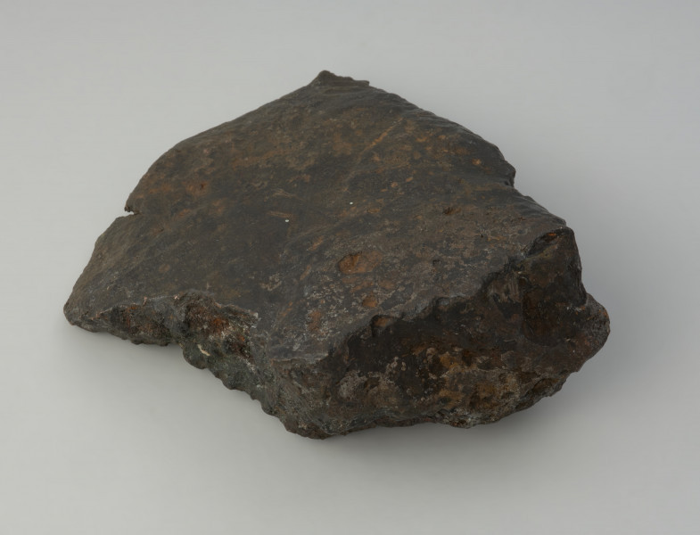 bryła metalu - Ujęcie w poziomie od tyłu. Fragment placka odlewniczego o nieregularnym, czworobocznym kształcie z zachowanym jednym z pierwotnych boków o ostrej krawędzi. W przekroju poprzecznym trójkatny.