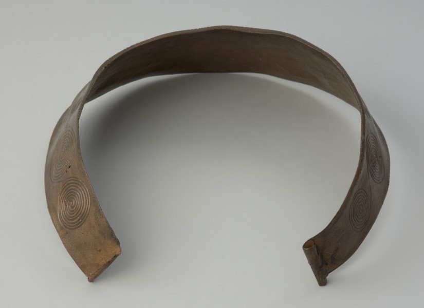 Brązowy naszyjnik kołnierzowaty - Ujęcie z tyłu. Brązowy naszyjnik kołnierzowaty jest zdobiony motywem  spiralnych pasm ułożonych w dwóch rzędach, oddzielonych podwójnym żeberkiem, ornamentowanym
drobnymi nacięciami i punktowaniem.