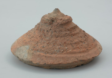 pokrywa gliniana - Ujęcie przodu w pionie. Pokrywkę wykonano z gliny na tarczy koła garncarskiego w technice całkowitego obtaczania. Ma stożkowaty kształt. Jej powierzchnia przyozdobiona jest motywami wielowątkowych zdobień takich jak dookolne linie ryte, linie faliste oraz ukośne nacięcia. Górną część pokrywki zwieńcza niewielki ozdobny guzek.