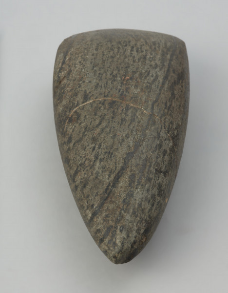 Siekiera kamienna - Ujęcie w pionie ostrzem w dół;  Siekiera kamienna o owalnym przekroju poprzecznym, ze spiczastym obuchem oraz z nieco asymetrycznym, łukowatym ostrzem. Powierzchnia jest silnie zeszlifowana, równa i gładka. Wykonana jest z surowca skalnego o zielonkawej barwie z czarnymi pasmami.