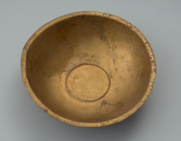 misa z rytem - Ujęcie z góry. Ozdobna misa wykonana została z jednego fragmentu blachy. Jej wąski, na zewnątrz wywinięty brzeg, miejscami opuszczony ku dołowi, ma zaokrągloną i lekko pogrubioną krawędź. Misa jest częściowo uszkodzona (jeden znaczny ubytek oraz kilkanaście mniejszych). Brakuje części ścianki od dna aż do wylewu. Pomimo ubytku i zniszczenia powierzchni zachowała się dekoracja ryta obejmująca wnętrze naczynia. Na nieco wklęsłym dnie widoczny jest duży medalion, z dodatkowo wydzieloną za pomocą podwójnej linii rytej częścią centralną wypełnioną liniami łamanymi. Na wewnętrznej powierzchni ścianki umieszczono symetrycznie cztery owalne medaliony z podobnym motywem, a pomiędzy nimi cztery 