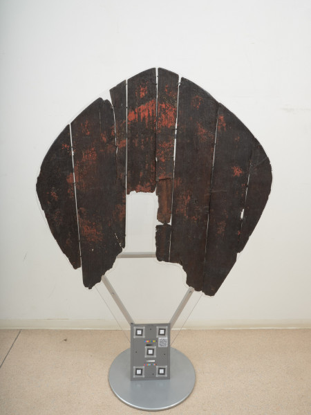 tarcza drewniana - Ujęcie z przodu. Tarcza drewniana, nawiązująca do tarcz typu migdałowatego. Wykonana została z sześciu deszczułek, zwanych też plankami, o szerokości 6,5–17 cm i grubości 0,4–0,7 cm, ściśle przylegających do siebie, spojonych najpewniej klejem pochodzenia roślinnego (żywicznego) lub zwierzęcego (kostnego). Zachowała się jedynie drewniana część tarczy oraz lewy uchwyt skórzany, tak zwany imacz, w postaci paska skóry o długości 42 cm, który przymocowany był za pomocą gwoździ. Po prawym imaczu zachowały się jedynie otwory, podobnie jak po pasie naramiennym. Frontową powierzchnię tarczy pokrywają ślady kolorowej dekoracji namalowanej bezpośrednio na deszczułkach czerwoną, żółtą i czarną farbą. Część centralną zajmuje czerwono-żółty stylizowany krzyż o rozszerzających się ramionach. Wzdłuż brzegu widnieją czerwone kółka i czarne owale z żółtymi rozetami wewnątrz, układające się naprzemiennie, oddzielone od części centralnej czarną kreską.