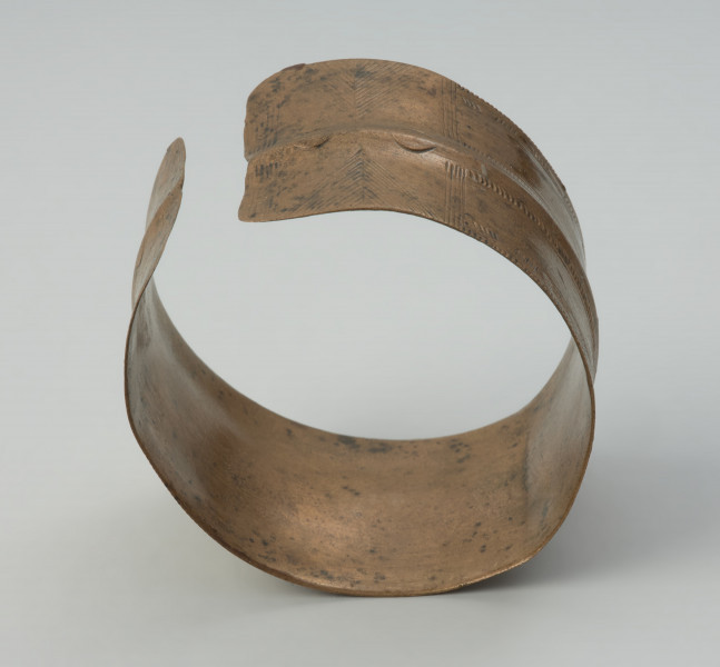 bransoleta taśmowata - Ujęcie w pionie. Bransoleta otwarta wykonana z wąskiej, dość grubej taśmy brązowej o przekroju płasko-wypukłym. Końce obręczy są ścięte prosto, a jej powierzchnię pokrywa ornament ryty złożony z grup ukośnych i pionowych kresek, poprzedzielanych motywem jodełki.