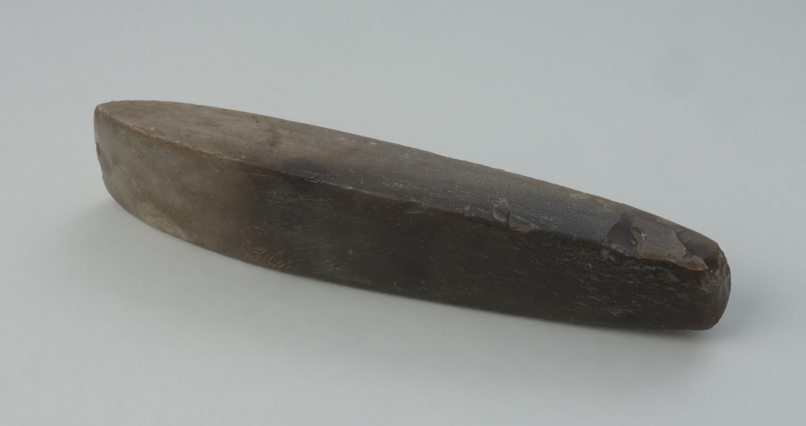 Dłuto krzemienne - Ujęcie z przodu skosem do tyłu. Dłuto krzemienne, czworościenne, trapezowate, o tępym obuchu. Powierzchnia szlifowana, przy ostrzu i w stronę obucha polerowana do połysku, a na obuchu negatywowa. Krzemień kredowy o barwie od jasno- do ciemnoszarej