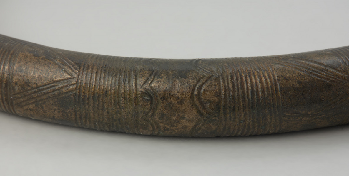 Brązowy naszyjnik - Powiększenie fragmentu naszyjnika z widocznymi zdobieniami. Brązowy naszyjnik wykonany z grubego okrągłego pręta. Jego zakończenia są odgięte na zewnątrz. Zdobiony jest poprzecznymi kreskami, trójkątami oraz łukami.