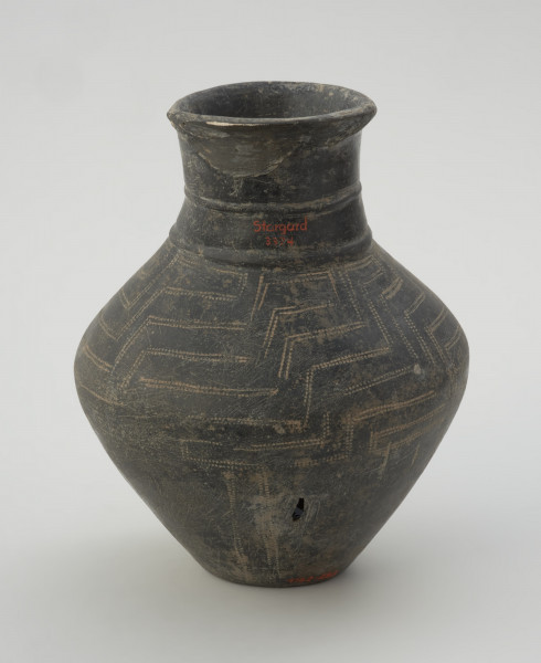 naczynie gliniane - Ujęcie z tyłu. Gliniane naczynie wąskootworowe o flaszowatej formie z wysoką cylindryczną szyjką. Powierzchnia jest wyświecana, barwy ciemnobrunatnej. Brzusiec jest dwustożkowaty, zdobiony techniką kółka zębatego motywem meandra schodkowego, a w dolnej partii pionowymi liniami. Szyjkę obiegają u podstawy dwie listy plastyczne.