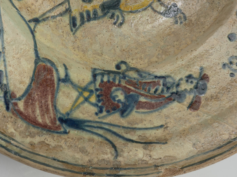 talerz - Ujęcie czwartego fragmentu. Wykonany na kole garncarskim z gliny o jasnej barwie ( z mała ilością tlenków żelaza). Dno słabo wyodrębnione przechodzi w niewysoki korpus o szerokim wlewie z wyraźnym kołnierzem. Szkliwo w tonacji żółtoszarej tylko na licowej powierzchni naczynia. Temat ornamentu wskazuje z dużym prawdopodobieństwem na to, że talerz był wyrobem jednostkowym wykonanym na zamówienie. Malatura przedstawia scenę rodzajowa: kobieta w niekompletnym stroju stoi pomiędzy dwoma mężczyznami. Jedna z tych postaci (większa) wyciąga ręce ku kobiecie , obok niej pies. Ponad postaciami , na kołnierzu, umieszczono napis: 