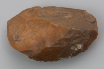 ciosak odłupkowy - ujęcie z przodu; Ciosak wykonany z odłupka krzemienia kredowego. Na obu bokach ciosaka zachowały się fragmenty oryginalnej strony wierzchniej i spodniej odłupka, z którego został wykonany. Przekrój soczewkowaty. Na bokach widoczne negatywy odbić kształtujących wykonanych miękkim tłukiem. Ostrze uformowane 3 odbiciami. Zabytek pokryty jest pomarańczowo-czerwoną patyną będącą efektem depozycji w środowisku o wysokiej zawartości żelaza.