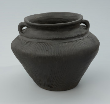 Waza gliniana barwy ciemnobrunatnej, wygładzana na powierzchni z uchem - Ujęcie z przodu. Waza gliniana barwy ciemnobrunatnej, wygładzana na powierzchni z uchem, które przechodzi w dookolne listwy plastyczne.