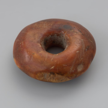 pacior bursztynowy - Ujęcie z boku. Pacior bursztynowy o pomarańczowo-brązowej barwie i dwustożkowatym kształcie ze ściętymi krawędziami. Powierzchnie są gładkie ze śladami obróbki, na boku znajduje się ubytek, kanalik umiejscowiono centrycznie. W jego wnętrzu widoczne są zatopione drobne listki lub płatki.