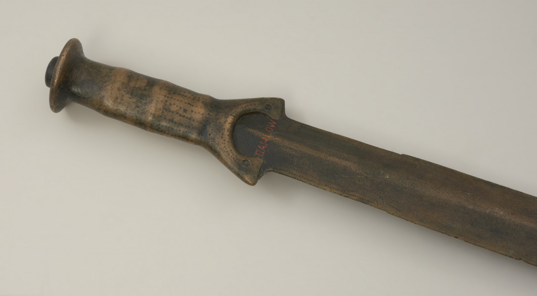 broń - Ujęcie rękojeści z tyłu w przybliżeniu. Brązowy miecz z rękojeścią zakończoną tarczką oraz szeroką głownią. Całość jest bogato zdobiona ornamentem punktowym oraz nacięciami.