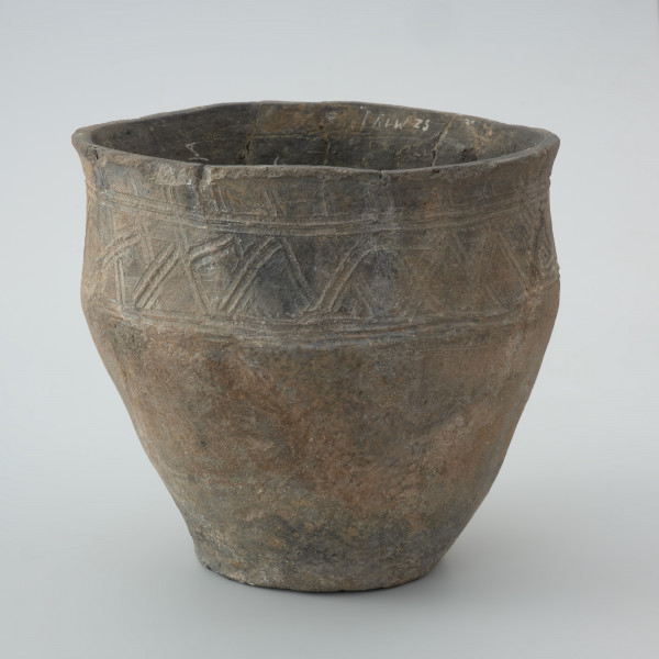 naczynie gliniane - Ujęcie przodu. Garnek ma kształt dwustożkowaty o esowatym profilu z charakterystyczną wychyloną na zewnątrz krawędzią wylewu oraz łagodny załom brzuśca znajdujący się w 2/3 wysokości. Na powierzchni pojemnika powyżej załomu występuje zdobienie w postaci krzyżujących się skośnych linii wpasowanych w podwójne, dookolne linie ryte. Naczynie wykonano w technice częściowego obtaczania na kole garncarskim. Uformowano je poprzez sukcesywne klejenie glinianych wałeczków albo taśm. W centralnej części dna znajduje się wklęsły odcisk po ośce, na której obracało się koło. Do produkcji wykorzystano masę garncarską z domieszką schudzającą drobno i średnioziarnistego tłucznia w postaci piasku i granitu.